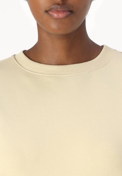 Ανοιχτό μπεζ φούτερ με στρογγυλή λαιμόκοψη, φτιαγμένο από υφή υφάσματος. Λεπτομέρεια με ριμπ λαιμό, απαλό ύφασμα και απλός σχεδιασμός χωρίς γραφικά ή σχέδια.