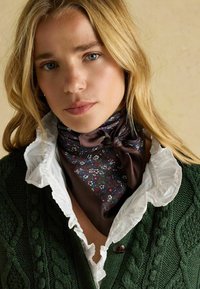 Sötétzöld, kötött kardigán texturált mintával, fehér fodros blúz fölé rétegezve. Nyak körül barna, virágmintás sál megkötve.