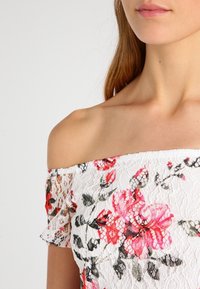 Bílý top bez ramínek z krajky s květinovými vzory v růžové, červené a zelené barvě. Obsahuje volánkové rukávy a jemný, texturovaný design.