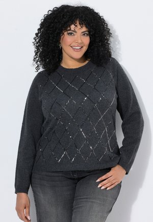 Vrouw met krullend zwart haar lacht en draagt een donkergrijze trui met een diamantvormig paillettenpatroon en donkergrijze spijkerbroek.
