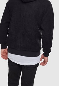 Felpa nera in pile con una superficie strutturata e soffice; presenta polsini e orlo a costine e una camicia bianca a strati sotto.