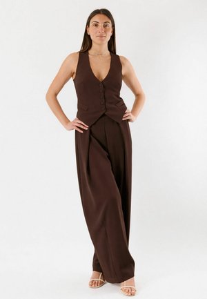 Donna in piedi con le mani sui fianchi che indossa un gilet marrone senza maniche con bottoni, pantaloni a gamba larga abbinati e sandali beige con punta aperta su sfondo bianco.