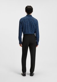 Long-sleeve shirt in blue with a textured, abstract pattern, paired with black trousers and shoes, viewed from the back.