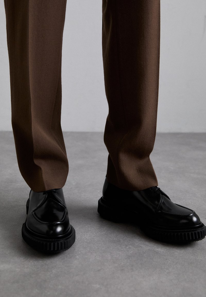 Schwarze Lederschuhe mit gerippten schwarzen Sohlen, kombiniert mit braunen, maßgeschneiderten Hosen aus einem glatten, strukturierten Stoff.