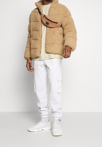Tan fleece pufferjakke, grå cargo sweatpants og beige sneakers. Brun corduroy crossbody taske båret over jakken. Casual ensemble.