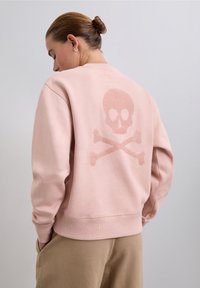 Kvinna med hår i knut som bär en rosa sweatshirt med en diskret glittrande dödskalle och tvärs över bena design på ryggen och beige byxor.