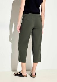 Pantalon court vert foncé avec une coupe décontractée, doté de deux poches arrière et d'une ceinture texturée. Associé à des sandales noires à glisser.
