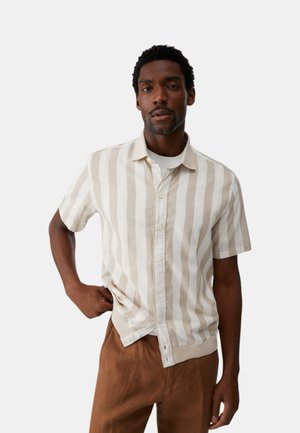 Uomo che indossa una camicia a righe verticali beige e bianche a maniche corte sopra una maglietta bianca e pantaloni marroni, in piedi con una mano sui fianchi.