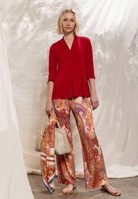 Haut rouge à manches longues avec un pli à l'avant, associé à un pantalon large à motifs cachemire colorés. Tenant un sac à main beige clair et une écharpe.