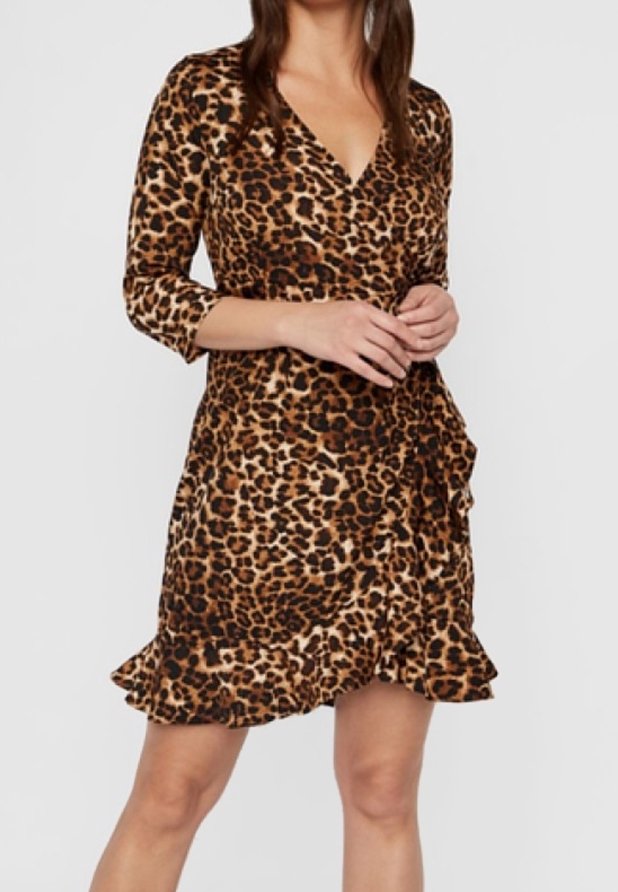 Femme portant une robe portefeuille marron et noire à imprimé léopard, manches trois-quarts et ourlet à volants, debout devant un fond uni.