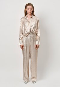 Metallic beige button-up shirt with a knotted front, paired with loose-fitting pants. Smooth texture and shiny finish. Pointed silver heels.