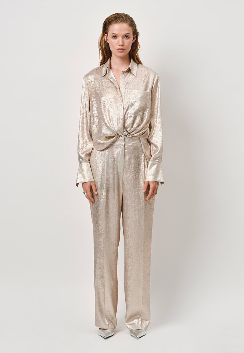Metallic beige button-up shirt with a knotted front, paired with loose-fitting pants. Smooth texture and shiny finish. Pointed silver heels.