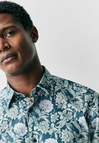 Bluse mit floralem Muster in dunkelblaugrün, mit detaillierten weißen und grünen Blumen, kurzer Kragen und durchgehender Knopfleiste. Glatte Stoffstruktur.