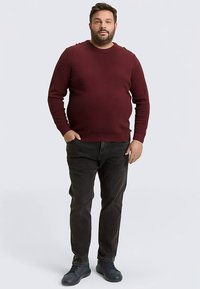 Bordeauxroter Pullover mit Rundhalsausschnitt und gerippter Textur, kombiniert mit dunkler Jeans und schwarzen Turnschuhen, steht vor einem einfachen Hintergrund.
