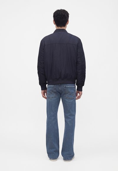 Homme debout face à l’opposé, portant une veste bomber noire et un jean bleu sur un fond clair uni.