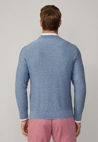 Gestrickter blauer Pullover mit strukturiertem Finish, Rundhalsausschnitt, langen Ärmeln und geripptem Saum. Über einem kragenlosen weißen Hemd getragen.