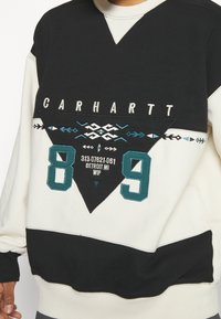 Schwarz-weißes Carhartt Sweatshirt mit geometrischen Mustern, großen Zahlen 8 und 9 sowie dem Text "313-07621-061 Detroit MI WIP" auf der Vorderseite.