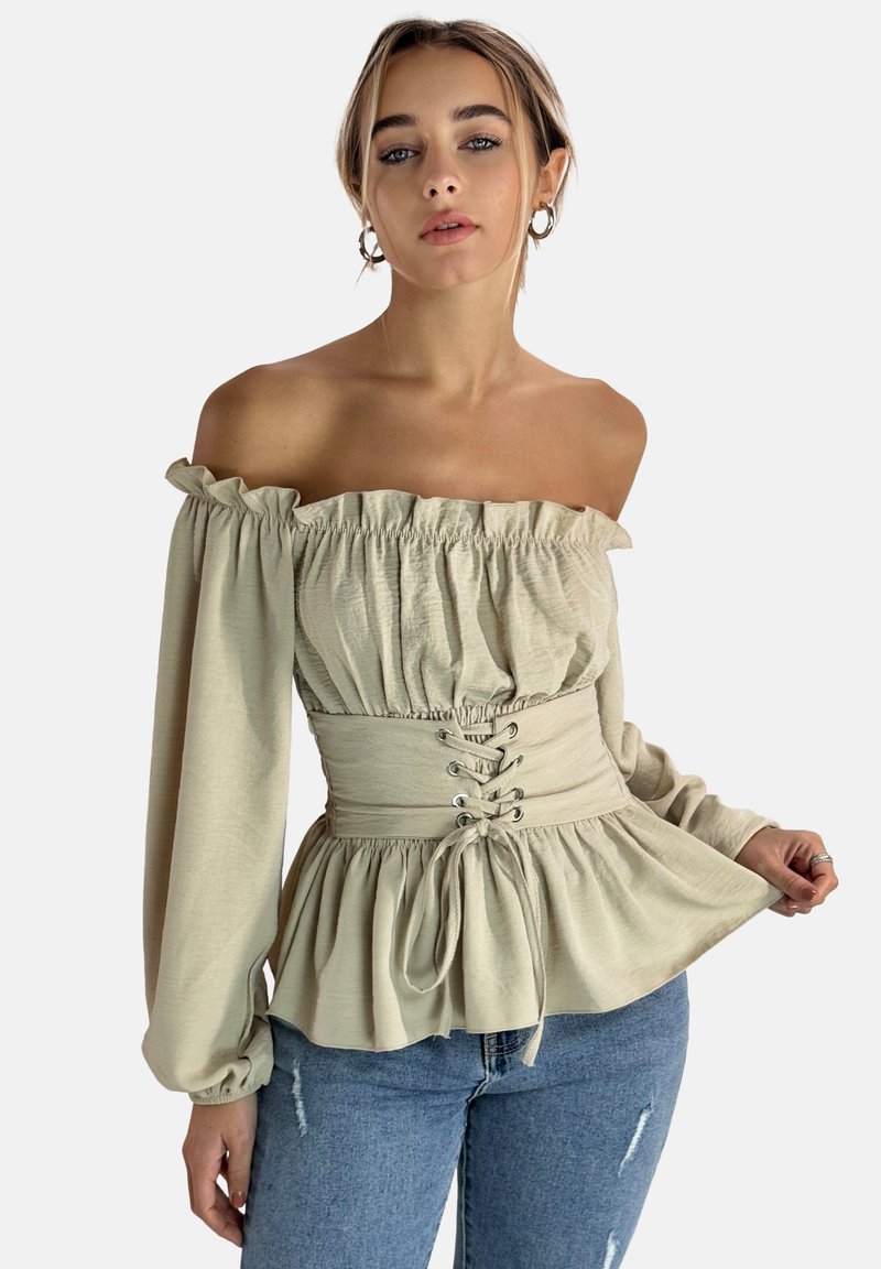 Beige Bluse mit schulterfreiem Schnitt und Rüschenausschnitt, tailliert mit Schnürdetail und fließenden Ärmeln, kombiniert mit blauen Jeans.