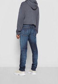 Blå denimjeans med slim fit, ljus blekning och distinkt sömnad på bakfickorna. Kombineras med en grå hoodie och ljusa sneakers.