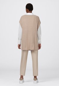 Beige, geripptes Strickwesten mit weiten Armlöchern, über einem weißen Hemd getragen. Kombiniert mit beigefarbenen Hosen und hellen Schuhen, in einem lässigen Schnitt.