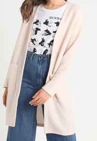 Frau trägt ein weißes T-Shirt mit Hand-Schattenspiel-Grafik und eine hellrosa offene Strickjacke über hoch taillierten blauen Jeans.