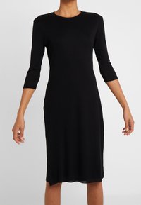 Robe noire, longueur genou, avec des manches trois-quarts et un col rond, fabriquée à partir d'un tissu lisse et léger avec une silhouette ajustée.