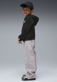 Felpa nera con cappuccio, jeans baggy di lavaggio chiaro e scarpe da ginnastica con design multicolore. Cappellino nero con logo, in piedi di lato su uno sfondo grigio.