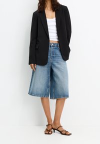 Blazer noir, crop top blanc et culottes en jean bleu clair. Blazer à ouverture avant, short à jambes larges avec ourlet effiloché, porté avec des sandales marron.