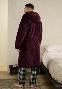 Mand står barfodet i soveværelset iført en bordeaux quiltet badekåbe med hætte og sort-hvide ternede pyjamasbukser.
