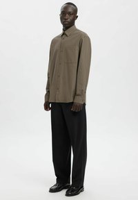 Brązowa koszula w kratę z długim rękawem, wykonana z miękkiej tkaniny, z kołnierzykiem zapinanym na guziki i kieszenią na piersi, zestawiona z luźnymi czarnymi spodniami.