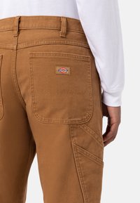 Pantalonii din pânză marron au două buzunare pe spate și o etichetă cu logo vizibil. Materialul are o suprafață texturată cu detalii de cusătură întărită.