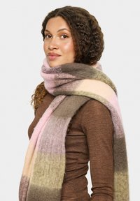 Ung kvinde med krøllet hår bærer en brun langærmet top og et tykt, blødt tørklæde med farveblokke i pink, beige og brun.