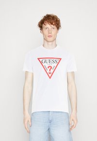 White cotton t-shirt featuring a red triangular logo design with "GUESS U.S.A." and "WASHED JEANS" text. Short sleeves and round neckline.