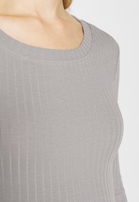 Maglia grigia a maniche lunghe con costine, caratterizzata da un collo rotondo e una texture morbida che mette in risalto linee verticali nel tessuto per un aspetto aderente.