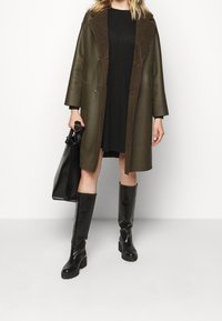 Cappotto in pelle verde oliva con colletto in shearling marrone, chiusura a doppio bottone, abbinato a un vestito nero e stivali neri al ginocchio, mentre tiene una borsa nera.