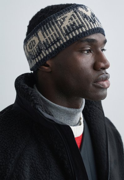 Homme à la peau foncée en profil portant un bonnet en tricot à motifs, une veste polaire noire et plusieurs pulls à col roulé superposés.