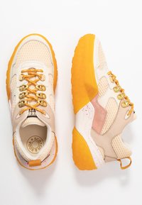 Beige Mesh- und Wildledersneaker mit leuchtend orangefarbenen Sohlen, orangefarbenen Schnürsenkeln, die durch goldene Ösen gezogen sind, und pinken Sprenkeln auf der weißen Zwischensohle.