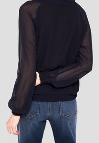 Blusa blu navy con maniche trasparenti, vita e polsini elasticizzati, e scollo rotondo; il tessuto è leggero e leggermente strutturato.