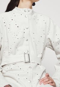 Robe-chemise en jean blanc avec détails perforés, boutons-pression, col mandarin et taille ceinturée. Texture lisse avec accents de ferrures argentées.