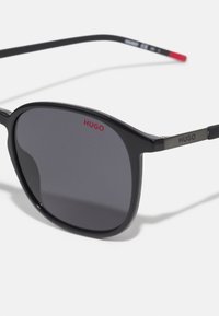 Ochelari de soare negri cu lentile gri, având un design cu cadru pătrat. Brațele afișează "HUGO" cu accente roșii și argintii.