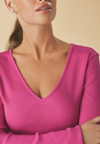 Tætsiddende langærmet top i ribbet varm pink stof. V-hals og brede manchetter fremhæver designet. Glat tekstur med let glans.