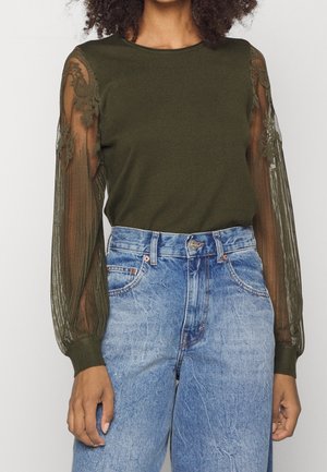 Femme portant un haut vert olive avec des manches longues en dentelle florale transparente, associé à un jean en denim bleu clair taille haute.
