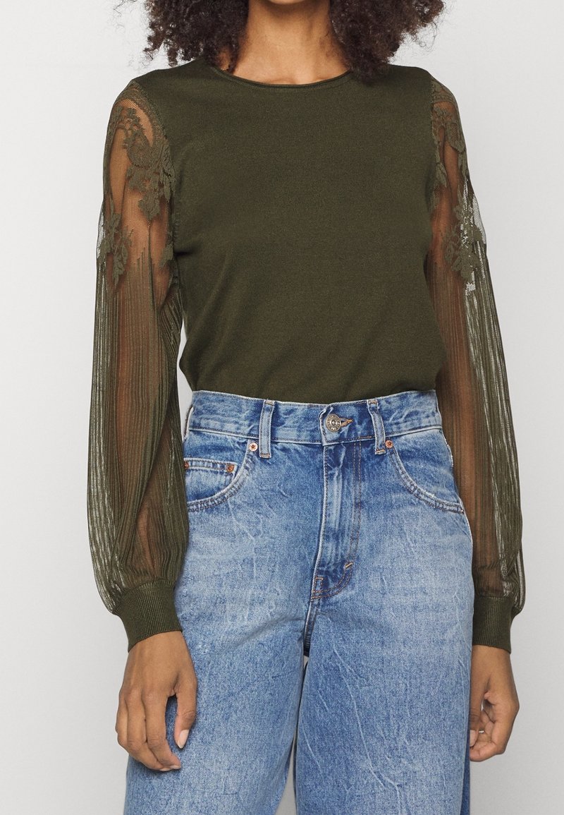Femme portant un haut vert olive avec des manches longues en dentelle florale transparente, associé à un jean en denim bleu clair taille haute.