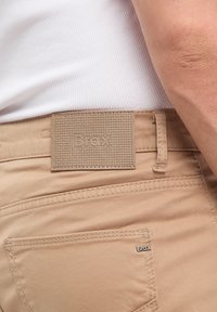 Chino beige chiaro con etichetta in tessuto testurizzato che mostra la scritta "Brax." Le caratteristiche includono cuciture visibili e un dettaglio della tasca posteriore.