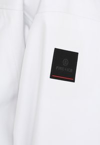 Tissu de veste blanche avec une texture lisse, présentant un patch carré noir portant l'inscription "FIRE+ICE" dans un coin et une ligne d'accent rouge.