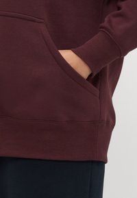 Sweatshirt bordeaux com um bolso canguru, punhos e bainha canelados, apresentado contra um fundo escuro. Tecido suave e texturizado.