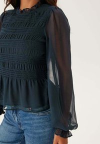 Tmavě zelená blůza se smockovanou spodní částí, průsvitnými dlouhými rukávy a volánkovými manžetami. Texturovaná látka s vrstveným designem a lehkým, splývavým vzhledem.