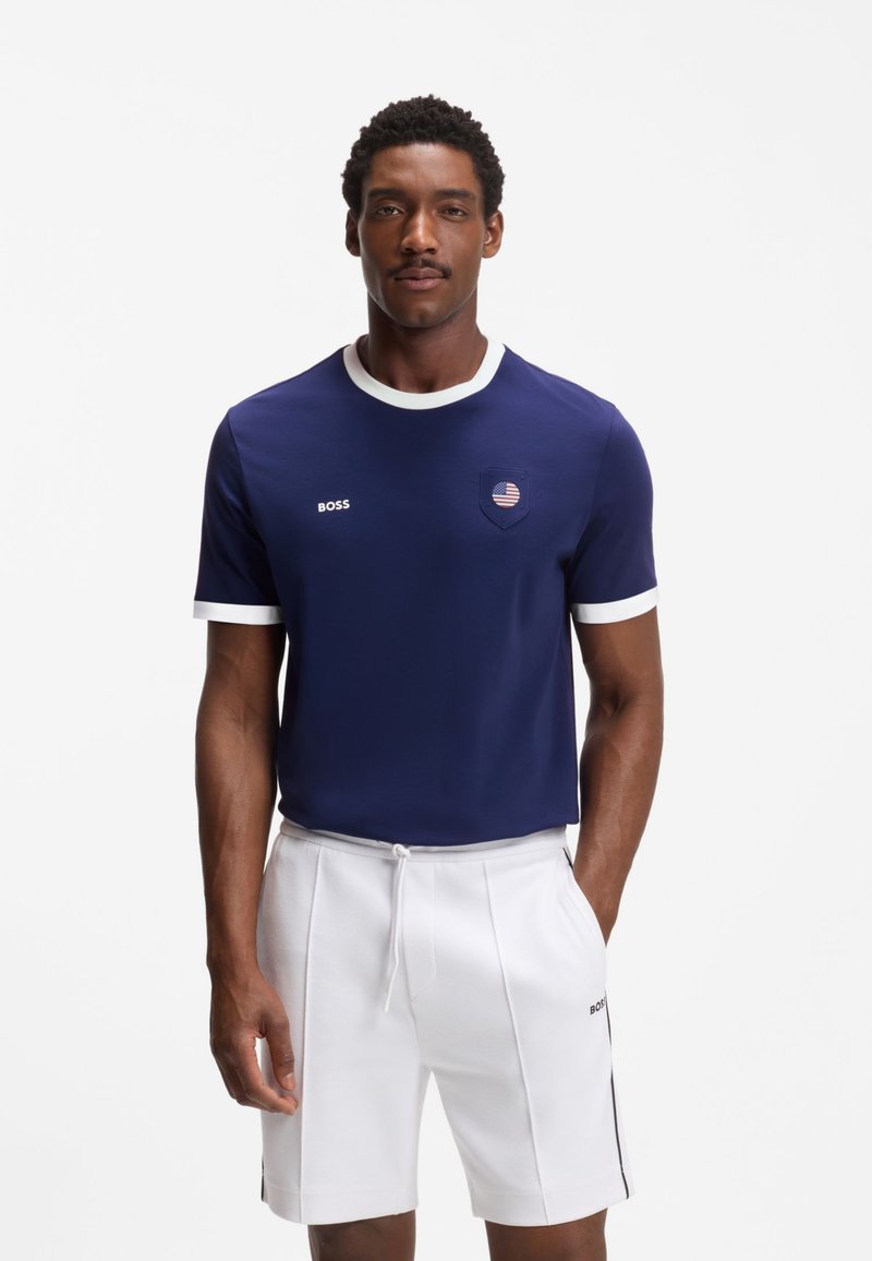 Homme debout portant un t-shirt bleu marine avec bordure blanche et un petit emblème, associé à un short blanc à cordon avec des bandes latérales.
