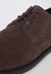 Chaussure en daim brun foncé à lacets avec une couture soignée et une semelle noire, présentée en gros plan sur un fond gris clair.