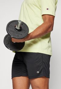 Halteres pretos segurados à mão, vestindo uma camisa de mangas curtas verde claro e calções pretos texturizados com um logótipo.